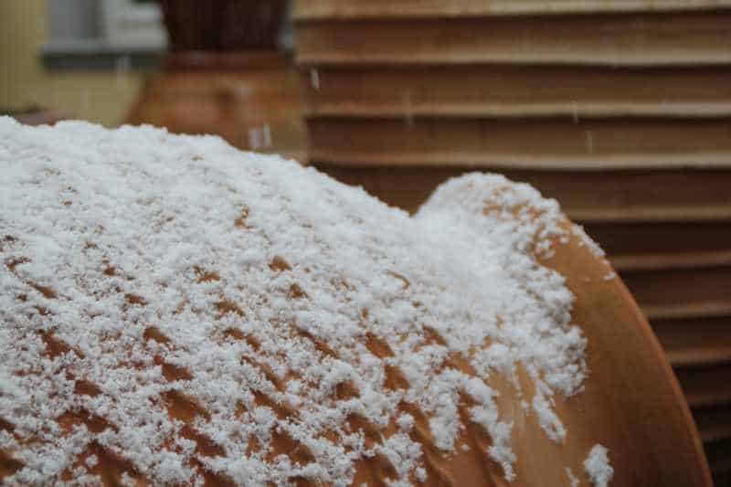 Nahaufnahme eines großen Terrakotta-Topfes (Terracotta-Töpfe), der teilweise mit einer Schneeschicht bedeckt ist. Im Hintergrund sind weitere Töpfe zu sehen. Schneeflocken fallen sanft und verstärken die Schneeschicht auf den Töpfen bei diesem Winterfest.