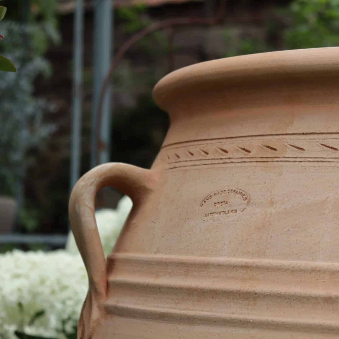 Eine große, hellbraune XXL-Terrakotta-Amphore „Thymus“ mit 100 cm Höhe, geschwungenem Henkel und eingravierten Zierlinien steht im Freien. Auf der Seite des Topfes befindet sich ein Stempel mit schwachem Text. Der Hintergrund zeigt üppiges grünes Laub und