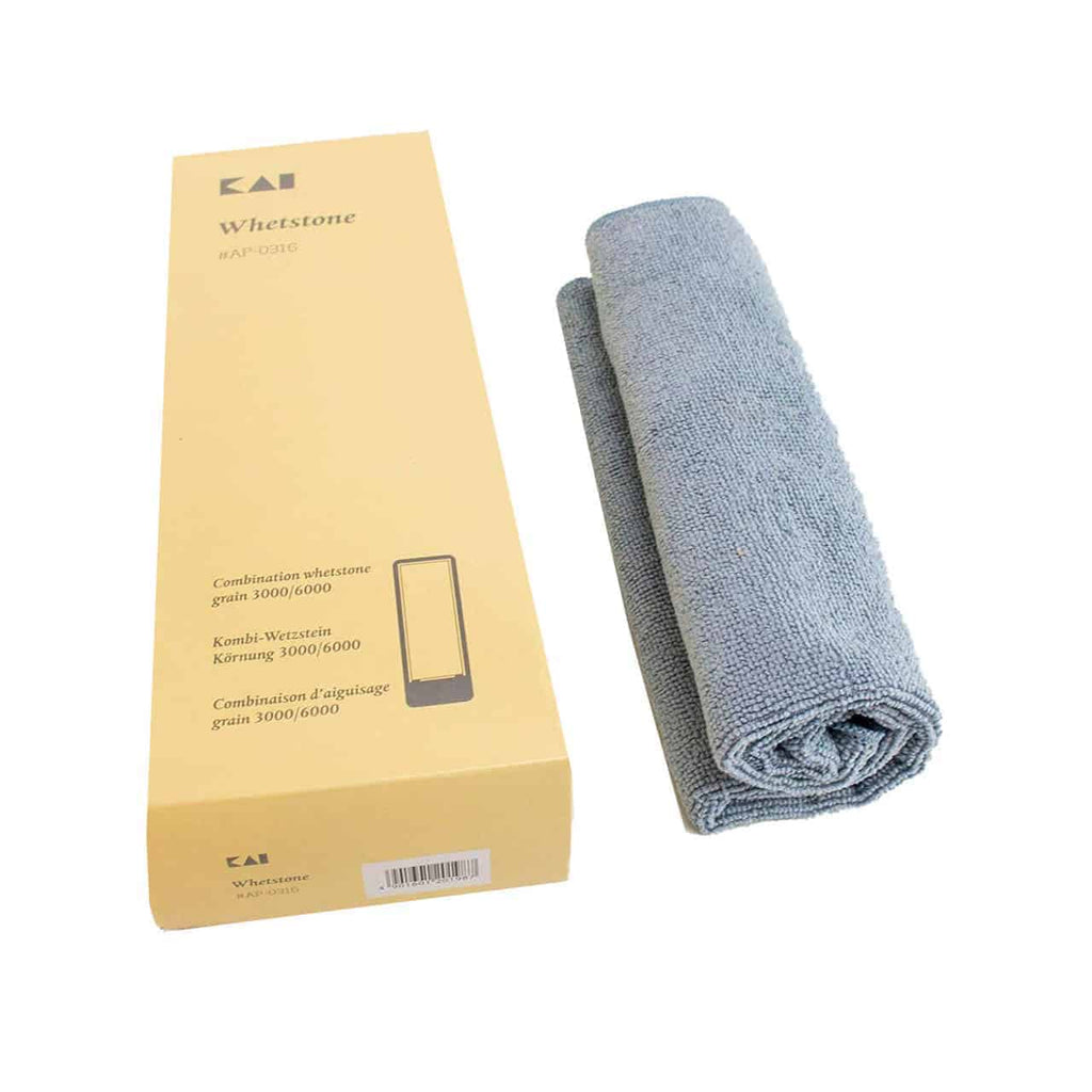 Links ist ein verpackter Kai AP-0316 | Schleifstein, Wetzstein 3000/6000 Körnung + Poliertuch zu sehen. Neben dem Karton liegt ein ordentlich gefaltetes graues Tuch.