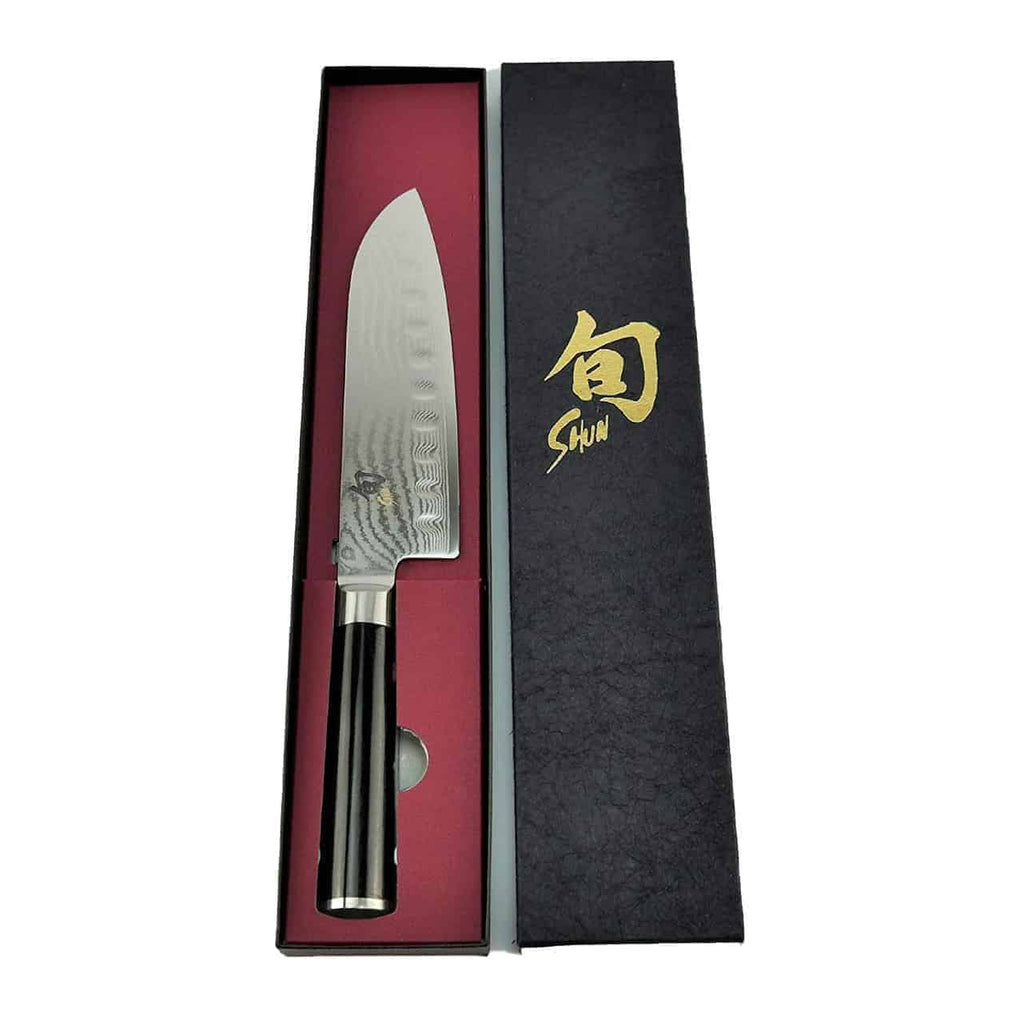 Ein Kai Shun Classic Santoku DM-0718 mit Kullenschliff, mit einer 18 cm langen polierten Klinge und einem schwarzen Griff, wird in einer schwarz-roten Box präsentiert, die mit goldenen japanischen Schriftzeichen und dem Wort „Shun“ verziert ist. Zum Angebot gehört auch ein Fassholzbrett mit den Maßen 34x21cm.