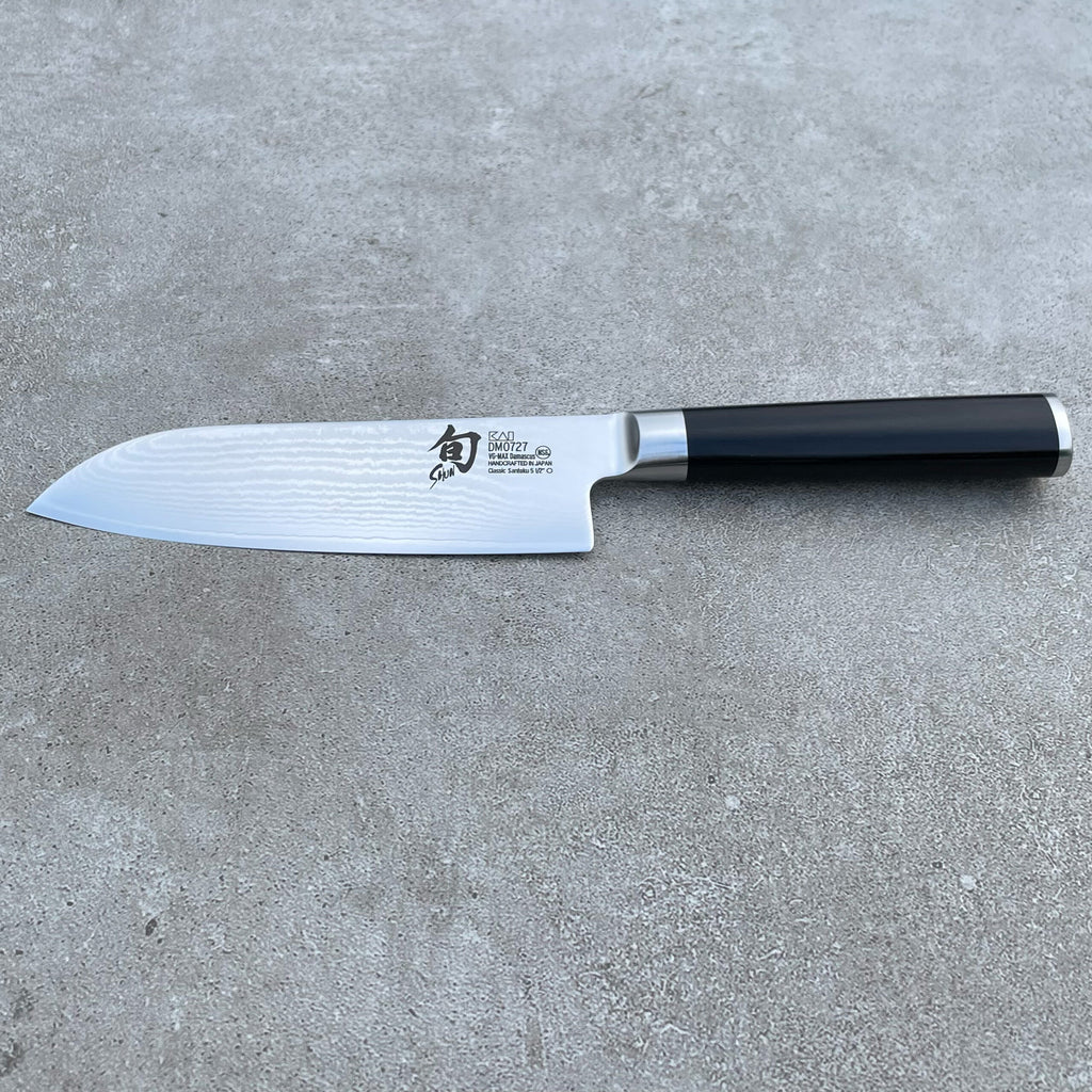 Ein elegantes Kai Shun Classic Santokumesser mit schwarzem Griff und glänzender, gemusterter Klinge liegt flach auf einer strukturierten grauen Oberfläche. Die ultrascharfe 14 cm lange Klinge aus dem DM-0727-Set zeigt aufwendig geätzte Markendetails.