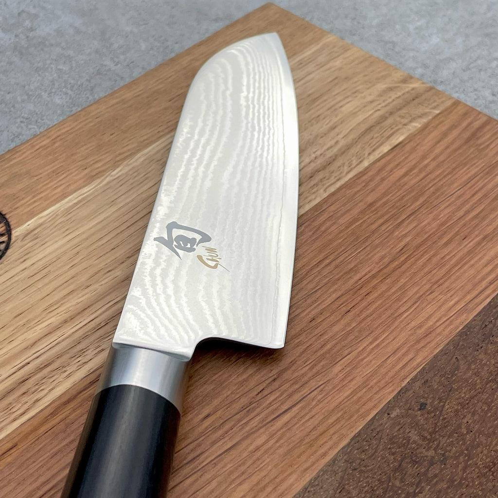 Eine Nahaufnahme des Kai Shun Classic Angebotsset | DM-0727 zeigt das ultrascharfe Santokumesser (14 cm) mit gewelltem Damastmuster und Logo, das auf dem massiven Eiche Schneidebrett (30x18 cm) ruht.