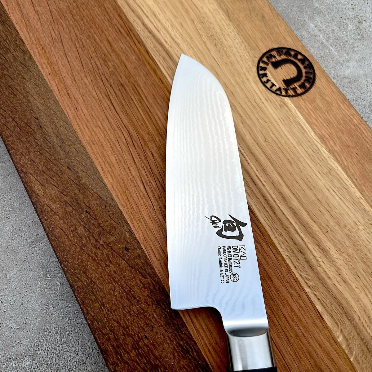 Ein Kai Shun Classic Angebotsset DM-0727 Santokumesser (14 cm) mit gemusterter Klinge liegt auf einem massiven Eichenholz-Schneidebrett (30x18 cm), beide zeigen hochwertige Handwerkskunst und das Markenlogo in der oberen rechten Ecke.