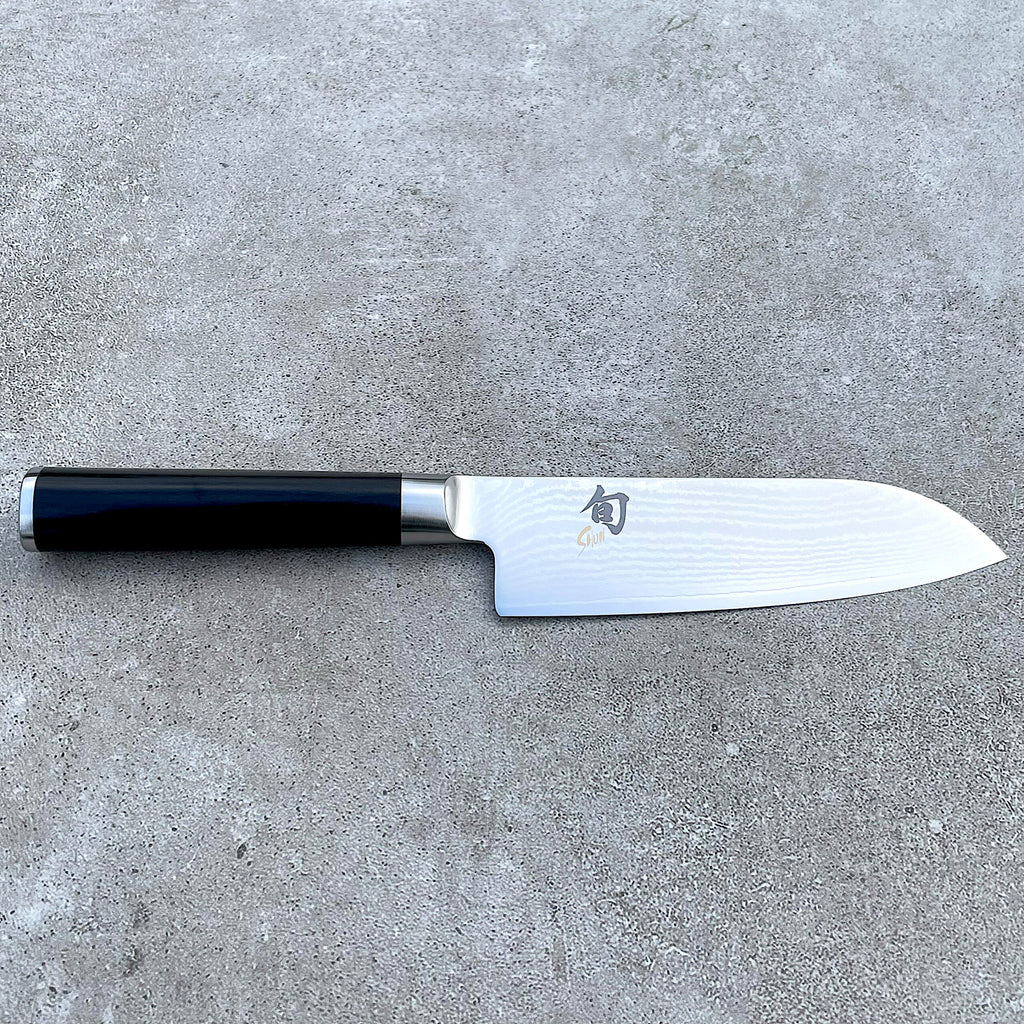 Ein Kai Shun Classic Santokumesser aus dem Angebotsset, ausgestattet mit einer 14 cm langen ultrascharfen Edelstahlklinge mit sichtbarer Musterung und schwarzem Griff, ruht horizontal auf einer grauen, strukturierten Oberfläche. Die Klinge ist mit japanischen Schriftzeichen und einem Logo in Griffnähe verziert, was es zu einem idealen Begleiter für das im Set enthaltene massive Eichen-Schneidebrett (30x18 cm) macht.