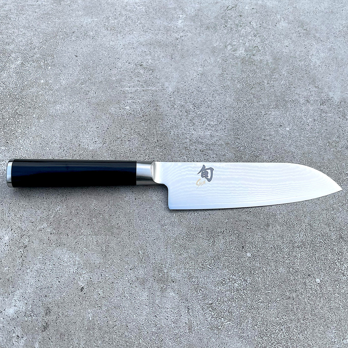 Ein Kai Shun Classic Santokumesser aus dem Angebotsset, ausgestattet mit einer 14 cm langen ultrascharfen Edelstahlklinge mit sichtbarer Musterung und schwarzem Griff, ruht horizontal auf einer grauen, strukturierten Oberfläche. Die Klinge ist mit japanischen Schriftzeichen und einem Logo in Griffnähe verziert, was es zu einem idealen Begleiter für das im Set enthaltene massive Eichen-Schneidebrett (30x18 cm) macht.