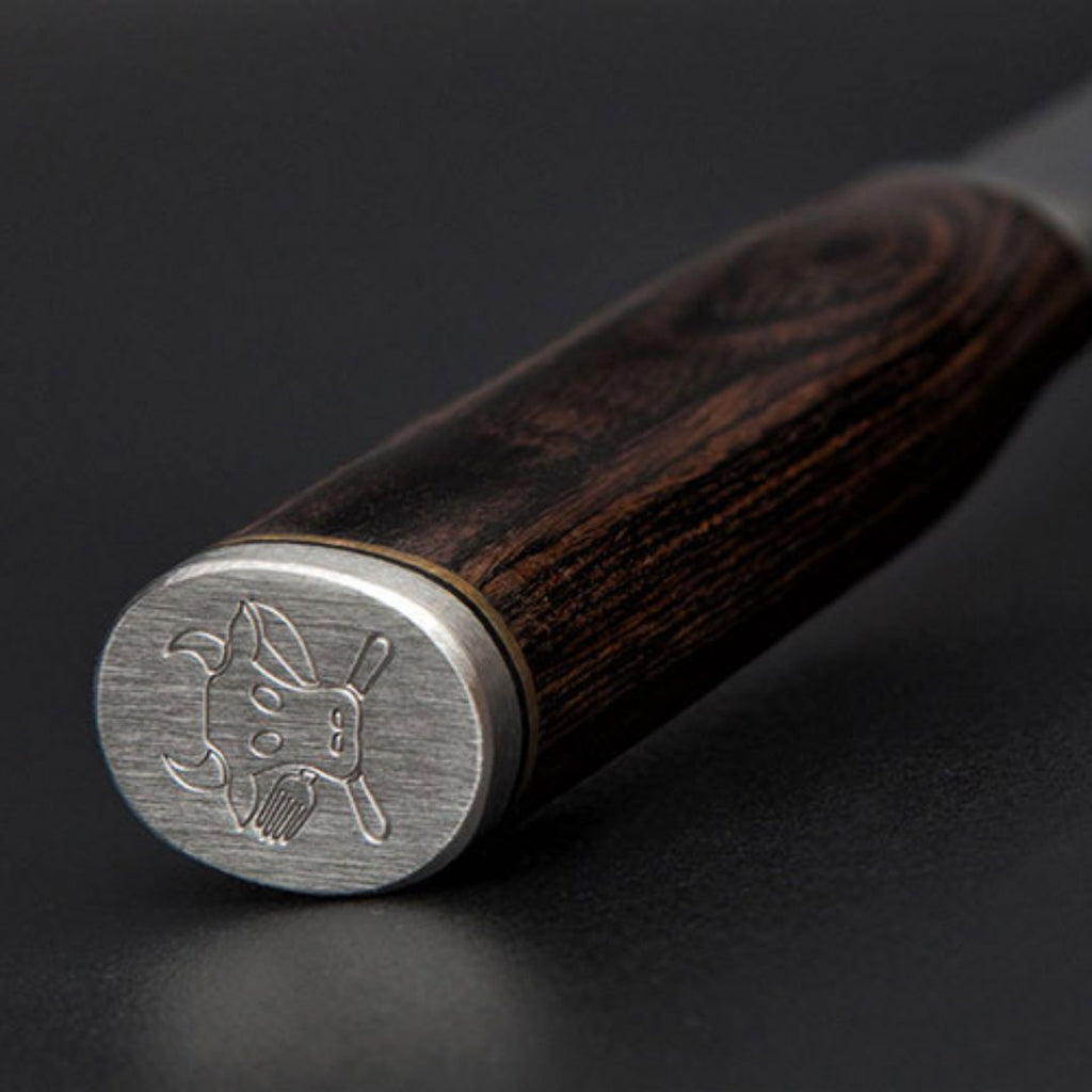 Nahaufnahme des Griffs des Kai Shun Tim Mälzer Santoku TDM-1702, mit dunklem Holzfinish und einer Endkappe aus Metall, auf der ein Stierkopf und gekreuzte Baseballschläger eingraviert sind. Klinge 18 cm, Edition "THE MAGIC KAI WISHLIST".