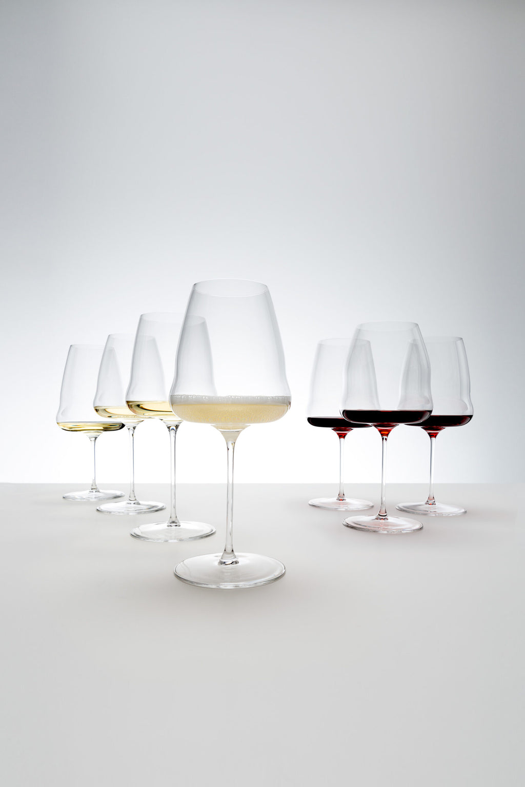 Sechs Weingläser, darunter das Champagnerglas Riedel Winewings mit Poliertuch in Geschenkverpackung, sind mit verschiedenen Weinsorten auf einer weißen Oberfläche gefüllt. Die Weine reichen in der Farbe von Hellgelb bis Dunkelrot. Der Hintergrund weist einen sanften Farbverlauf aus Weiß und Hellgrau auf, der dem Bild ein klares und minimalistisches Aussehen verleiht.