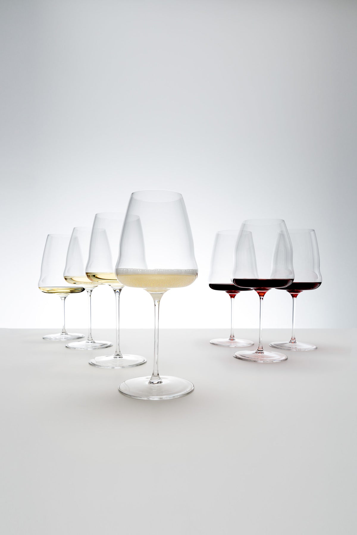 Sechs Weingläser, darunter das Champagnerglas Riedel Winewings mit Poliertuch in Geschenkverpackung, sind mit verschiedenen Weinsorten auf einer weißen Oberfläche gefüllt. Die Weine reichen in der Farbe von Hellgelb bis Dunkelrot. Der Hintergrund weist einen sanften Farbverlauf aus Weiß und Hellgrau auf, der dem Bild ein klares und minimalistisches Aussehen verleiht.