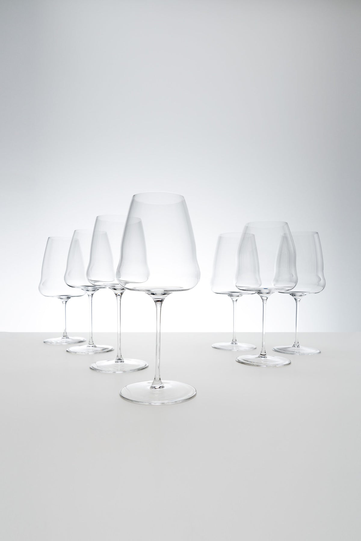 Ein Set aus sechs makellosen Champagnergläsern von Riedel Winewings, angeordnet in zwei Reihen auf einer glatten weißen Oberfläche vor einem hellgrauen Hintergrund. Die Gläser sind sauber, klar und elegant gestaltet und reflektieren das Licht subtil. Perfekt, um jeder Zusammenkunft einen Hauch von Raffinesse zu verleihen oder als Teil einer exquisiten Geschenkverpackung, komplett mit einem Poliertuch.