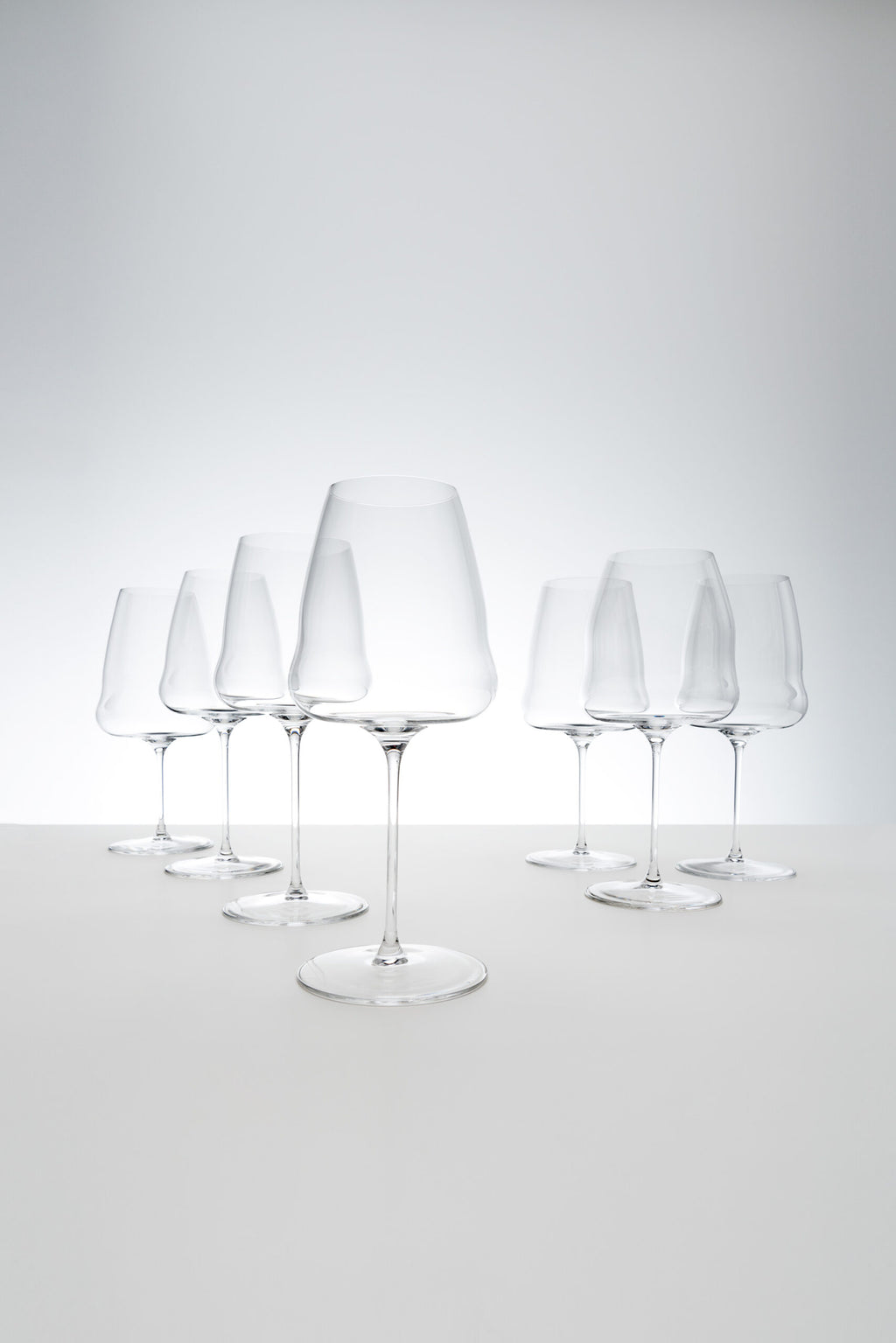Ein Set aus sechs makellosen Champagnergläsern von Riedel Winewings, angeordnet in zwei Reihen auf einer glatten weißen Oberfläche vor einem hellgrauen Hintergrund. Die Gläser sind sauber, klar und elegant gestaltet und reflektieren das Licht subtil. Perfekt, um jeder Zusammenkunft einen Hauch von Raffinesse zu verleihen oder als Teil einer exquisiten Geschenkverpackung, komplett mit einem Poliertuch.