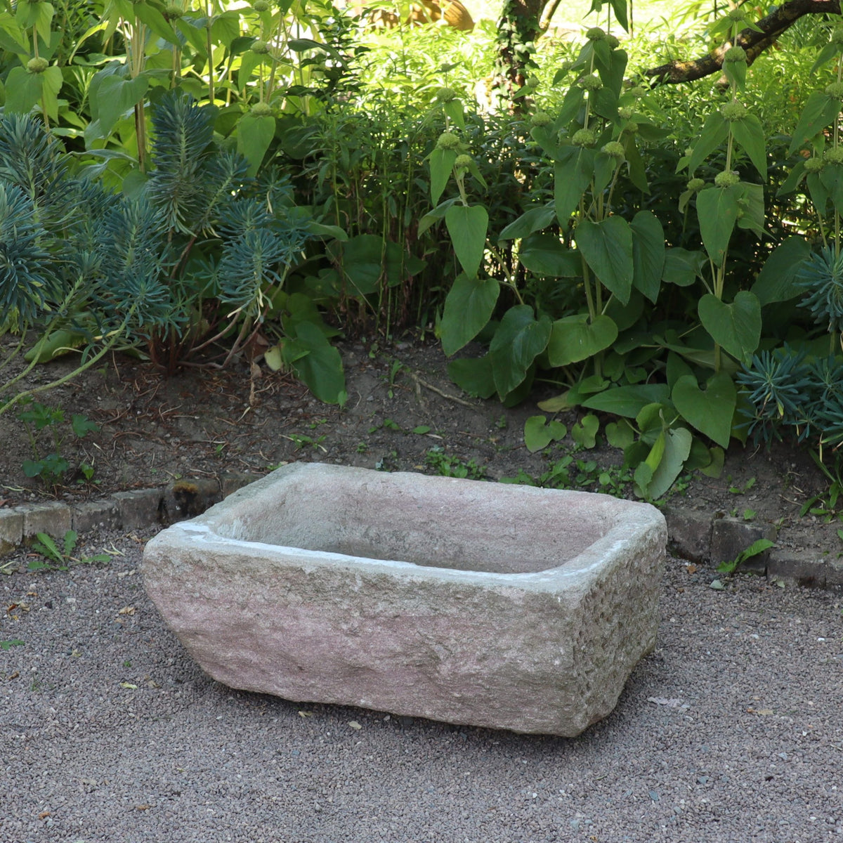 Der nostalgische Pflanzkübel aus Sandstein (80 x 59 x 29 cm) steht auf Kies vor dichter grüner Bepflanzung im Garten.
