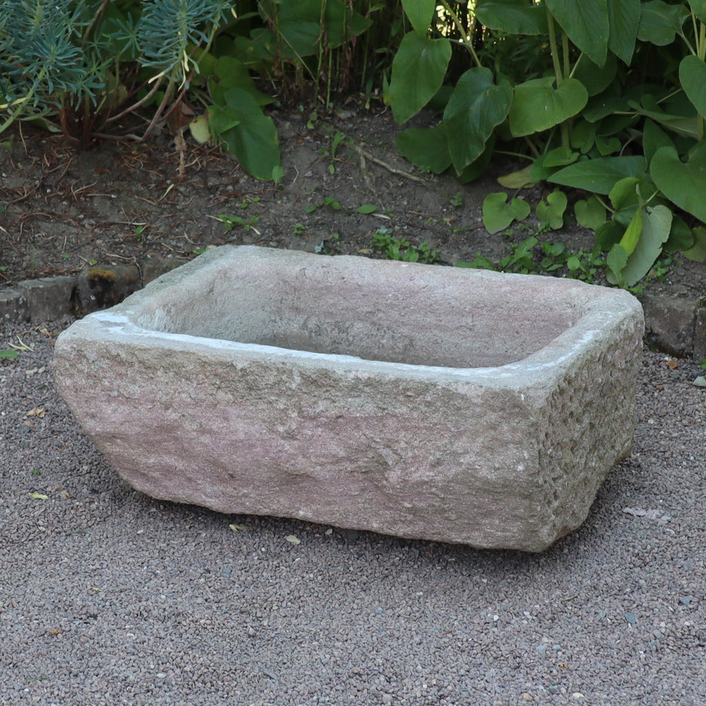 Ein nostalgischer Pflanzkübel aus Sandstein (80 x 59 x 29 cm) steht auf Kies und ist von grünen Blattpflanzen im Garten umgeben.