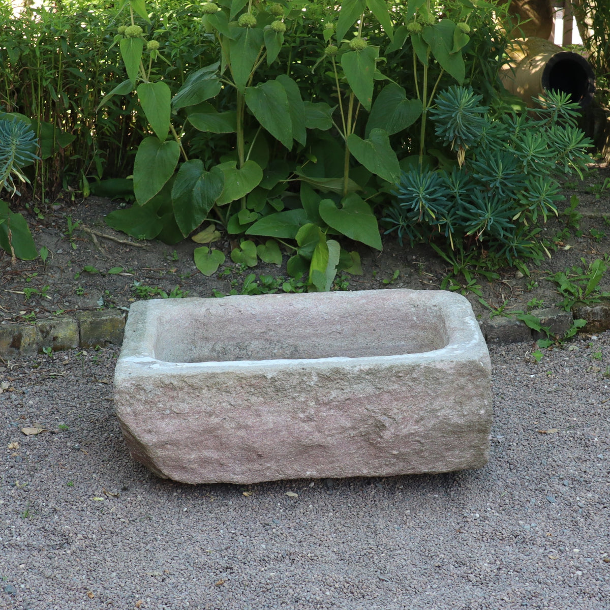 Ein nostalgischer Pflanzkübel aus Sandstein (80 x 59 x 29 cm) steht auf Kies und ist von grünen Pflanzen im Garten umgeben.