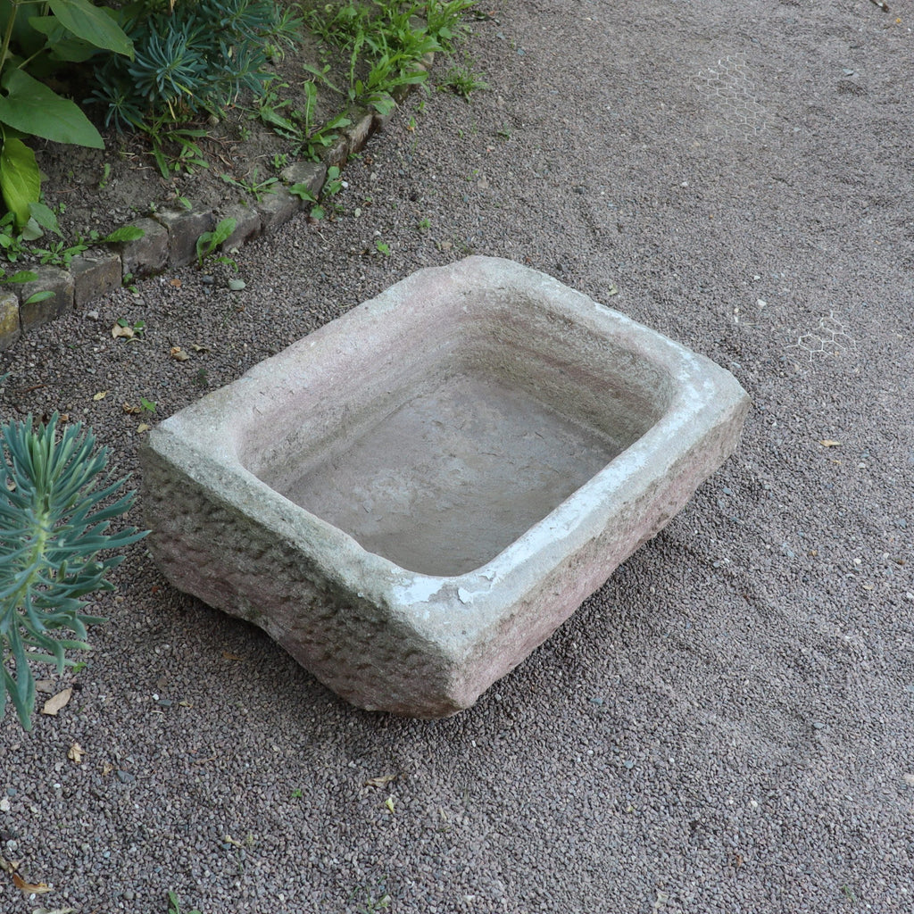 Der nostalgische Pflanzkübel aus Sandstein (80 x 59 x 29 cm) mit rustikalen Kanten steht auf Kieseln, umgeben von kleinen Steinen und grünen Pflanzen.