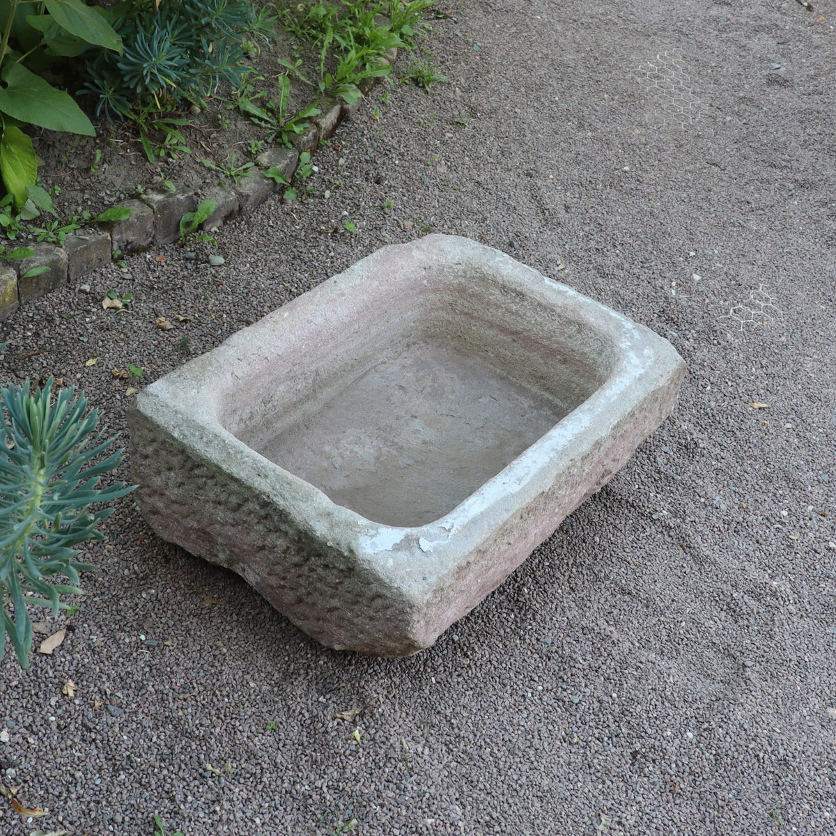 Der nostalgische Pflanzkübel aus Sandstein (80 x 59 x 29 cm) mit rustikalen Kanten steht auf Kieseln, umgeben von kleinen Steinen und grünen Pflanzen.