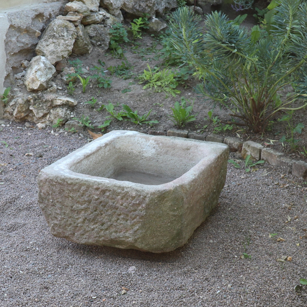Der nostalgische Pflanzkübel aus Sandstein (80 x 59 x 29 cm) steht auf Kies, umgeben von Steinen, Grünpflanzen und Erdflecken in einer Gartenanlage.