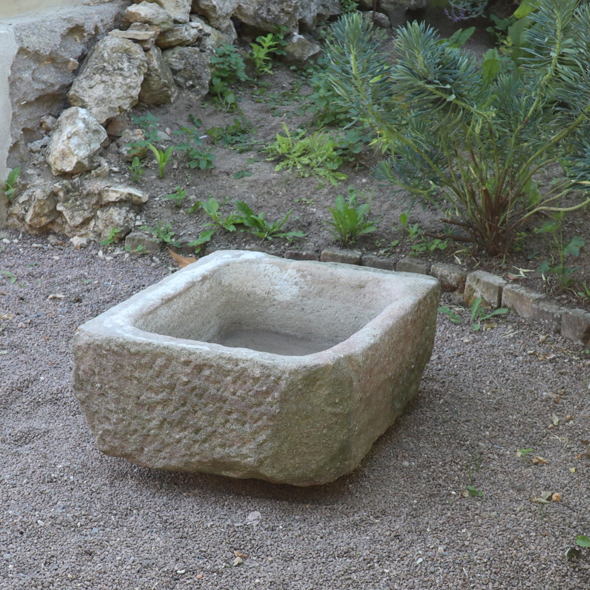 Der nostalgische Pflanzkübel aus Sandstein (80 x 59 x 29 cm) steht auf Kies, umgeben von Steinen, Grünpflanzen und Erdflecken in einer Gartenanlage.