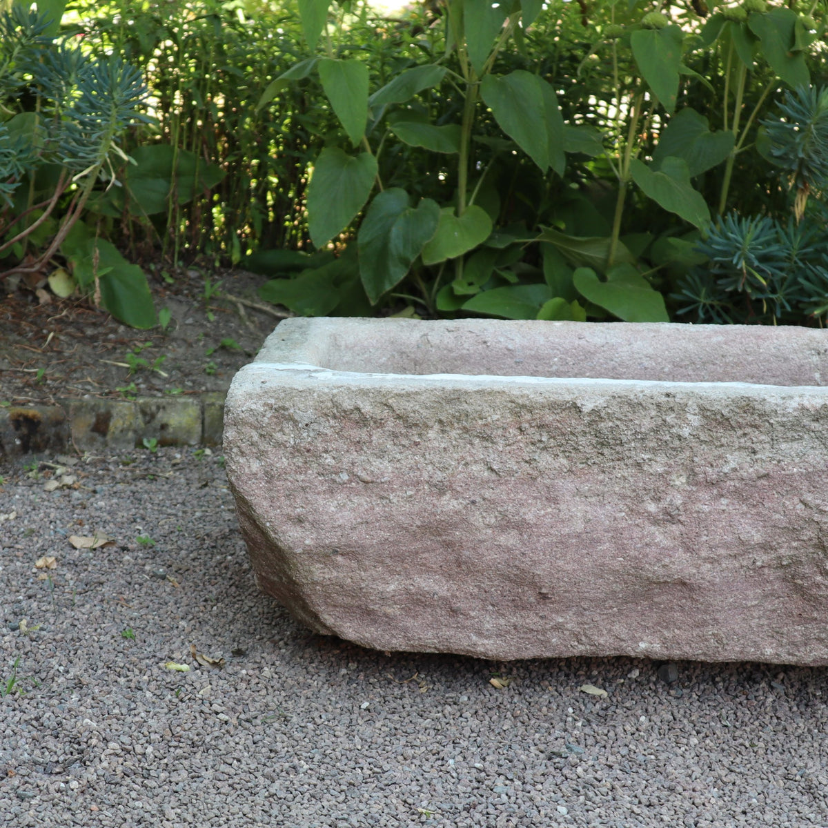 Der nostalgische Pflanzkübel aus Sandstein (80 x 59 x 29 cm) steht auf Kies, teilweise beschattet von Laub, mit Grünpflanzen und einem Gartenbeet im Hintergrund. Sein verwittertes Aussehen unterstreicht die natürliche Umgebung.