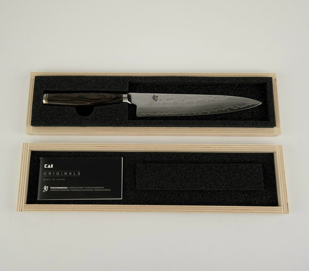 Ein Kochmesser mit gemusterter Klinge und dunklem Holzgriff wird in einer mit Schaumstoff ausgekleideten Holzbox präsentiert. Der geöffnete Deckel gibt den Blick frei auf das Kai Shun Tim Mälzer Angebotsset Allzweckmesser TDM1701, beiliegend eine Karte mit der Aufschrift „Kai Shun Tim Mälzer.