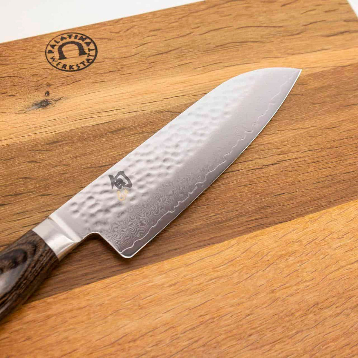 Ein hochwertiges Kai Shun Tim Mälzer TDM-1702 Santoku Messer, 18 cm lang und aus gehämmertem Damaststahl mit Holzgriff, ruht auf einem Küchenarbeitsbrett aus hellem und dunklem Holz. Das 40x30 cm große Schneidebrett weist in der oberen linken Ecke ein eingraviertes Logo auf, während das Messer ein Logo in der Nähe des Kropfes aufweist.