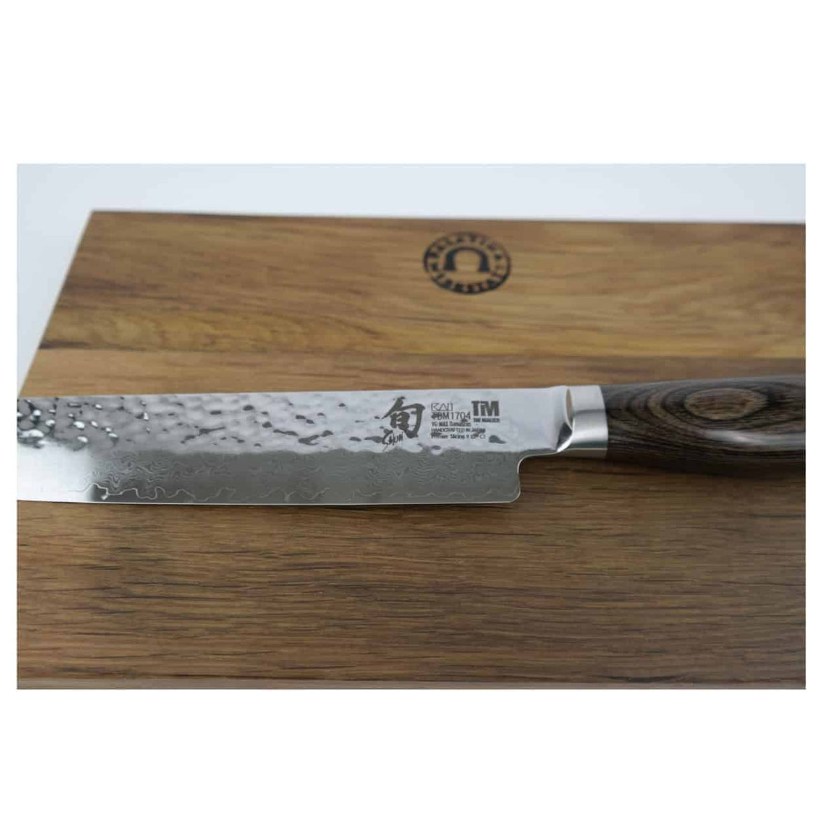 A Kai Shun Premier Tim Mälzer (TDM-1704) ultrascharfes japanisches Schinkenmesser mit einer gehämmerten Damaststahl-Klinge und einem dunklen Holzgriff liegt auf einem massiven Schneidebrett aus Fassholz (30x18 cm). Die Klinge zeigt eine kunstvolle Verarbeitung und weist nahe der Rückseite einige Markendetails auf.