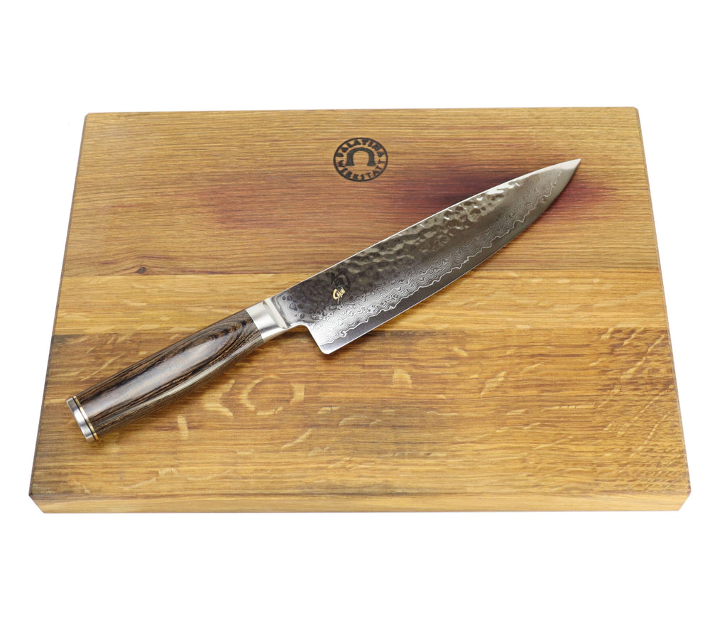 Ein Kai Shun Premier Tim Mälzer TDM-1706 Kochmesser mit einer 18 cm langen gemusterten Damaststahlklinge und einem Holzgriff ruht auf einem XXL-Schneidebrett mit den Maßen 40x30 cm. Das Schneidebrett, das am oberen Rand mit einem Logo verziert ist, steht auf einem weißen Hintergrund.