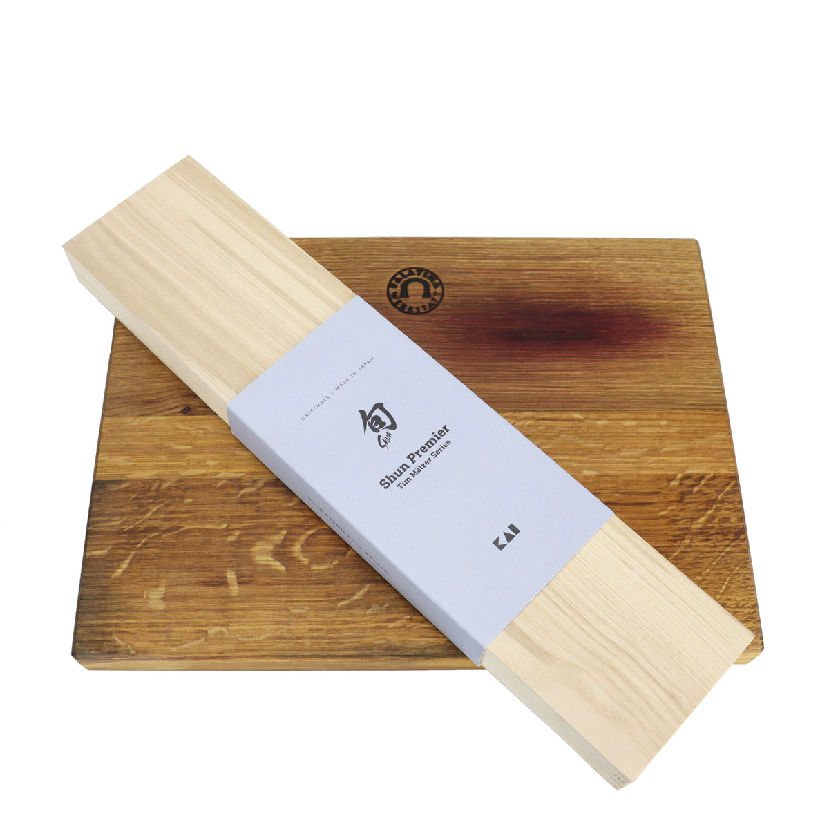 Das Kai Shun Premier Tim Mälzer TDM-1706 Kochmesser aus Damaststahl mit einer 18 cm langen Klinge wird wunderschön auf einem XXL-Schneidebrett aus Holz im Format 40 x 30 cm präsentiert. Der hölzerne Messerhalter ist in hellblaues Papier mit der Aufschrift „Kai Shun Premier“ eingewickelt und wird neben dem Messer ausgestellt. Beide Artikel stehen auf einem weißen Hintergrund, wobei das Schneidebrett an einer Kante eine Rille aufweist, um die exquisite Handwerkskunst des Damaststahlmessers elegant zur Geltung