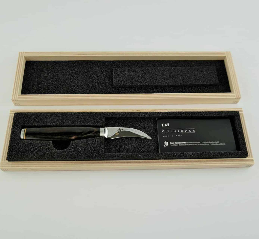 Eine hölzerne Geschenkbox mit einem Kai Shun Premier Tim Mälzer TDM-1715 Tourniermesser mit schwarzem Griff und silberner Klinge. Die Box hat eine Schaumstoffeinlage, die das 5,5 cm lange Messer sicher hält. Neben dem Messer befindet sich eine Visitenkarte mit Text, alles auf einem schlichten weißen Hintergrund. Zusätzlich enthält das Set ein klingenschonendes Schneidebrett aus Fasseiche in den Maßen 25x15 cm. Dieses exquisite Kai Shun Premier Geschenkset ist ein elegantes Geschenk.