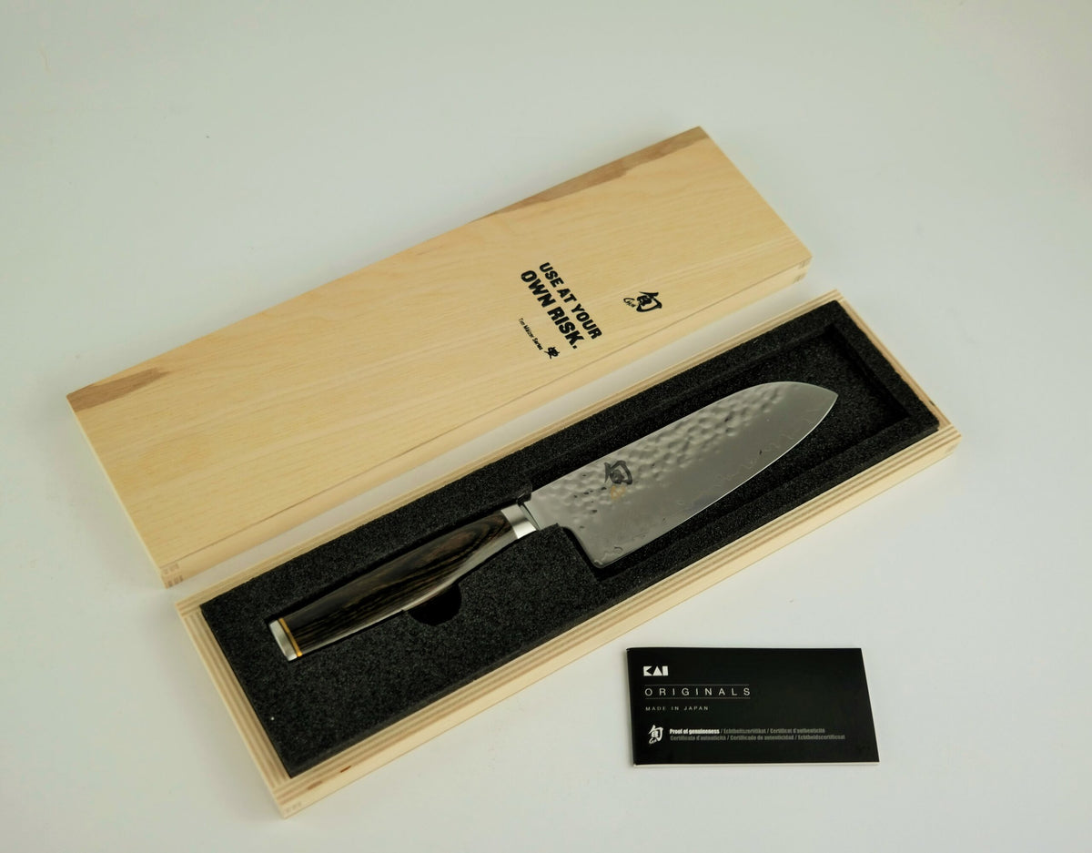 Das Kai Shun Premier Tim Mälzer TDM-1727 kleines Santoku mit 14 cm langer Klinge wird in einer offenen Holzbox geliefert, neben der sich der Deckel befindet. Das Messer mit dem schwarzen Griff liegt in schwarzem Schaumstoff, neben der Box befindet sich eine schwarze Karte mit weißem Text. Ein passendes Schneidebrett im Format 30 x 18 cm ist ebenfalls enthalten. Der Hintergrund ist eine schlichte weiße Oberfläche.