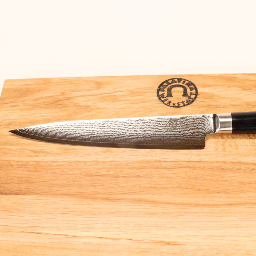 Ein Kai Shun DM-0701 Allzweckmesser (15 cm) mit dunklem Griff liegt auf einem 30x20 cm großen Schneidebrett aus Eichenholz, das in der Ecke ein rundes Logo trägt.