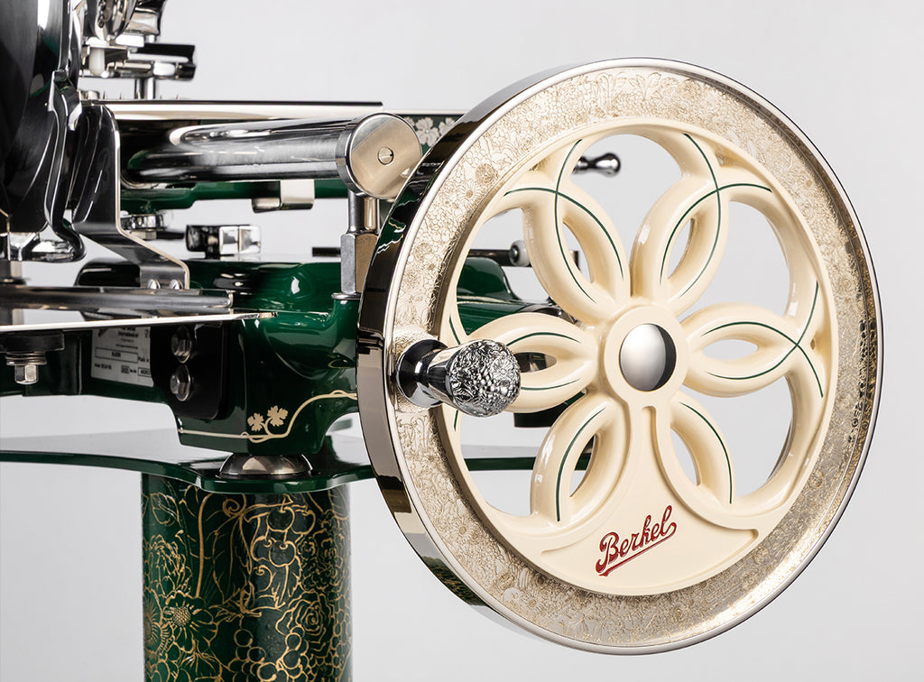Nahaufnahme der Berkel Volano B114 Sondermodell Schwungradmaschine inkl. Standfuß + Fassholzbrett. Standfuß + Fassholzbrett, im Vintage-Design mit cremefarbenem Handrad, verziert mit floralen Mustern und grünen Akzenten auf einem glänzend grünen Sockel.