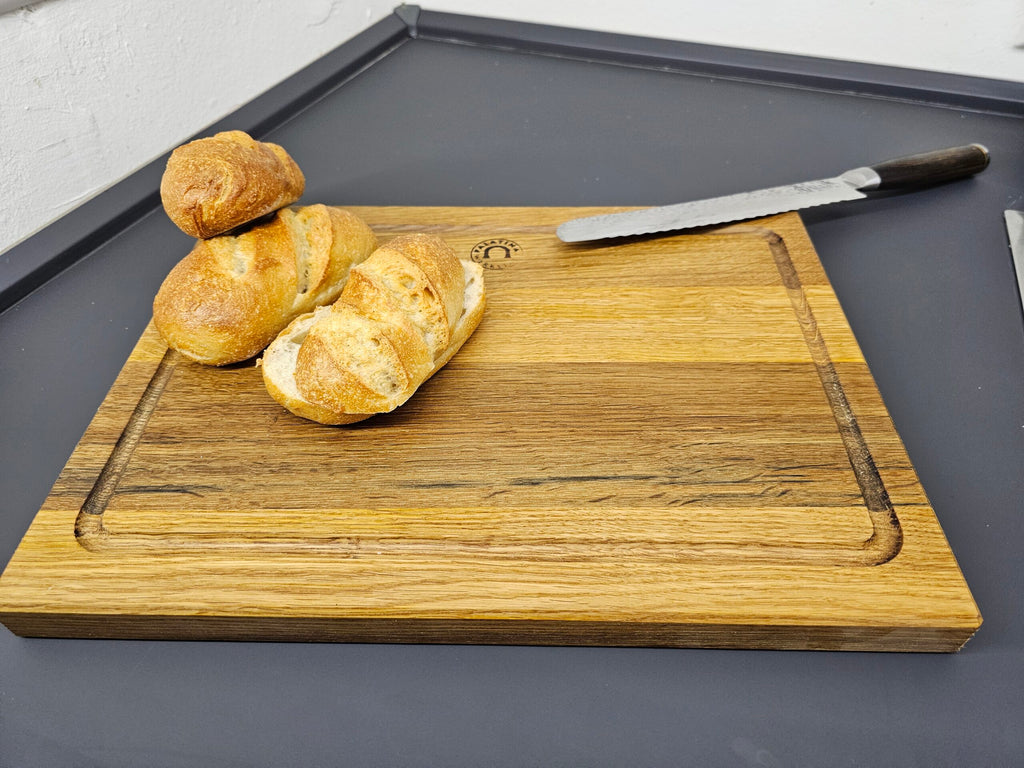 Drei Brotscheiben liegen auf einem hölzernen Palatina Küchenbrett mit Saftrille 40x30 cm aus Weinfassholz. Rechts neben dem Brot liegt ein gezacktes Messer mit dunklem Griff auf dem Schneidebrett. Das Schneidebrett liegt auf einer dunkelgrauen Oberfläche.