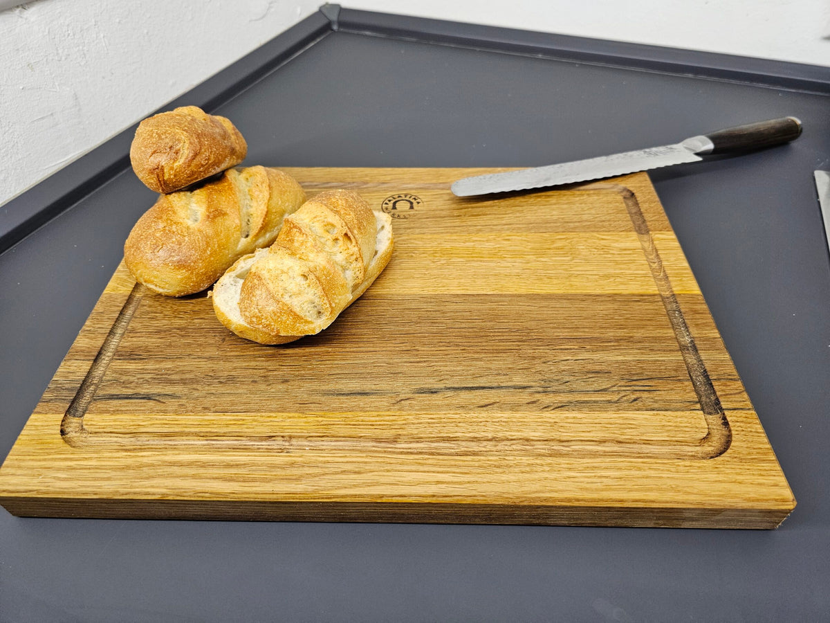 Drei Brotscheiben liegen auf einem hölzernen Palatina Küchenbrett mit Saftrille 40x30 cm aus Weinfassholz. Rechts neben dem Brot liegt ein gezacktes Messer mit dunklem Griff auf dem Schneidebrett. Das Schneidebrett liegt auf einer dunkelgrauen Oberfläche.