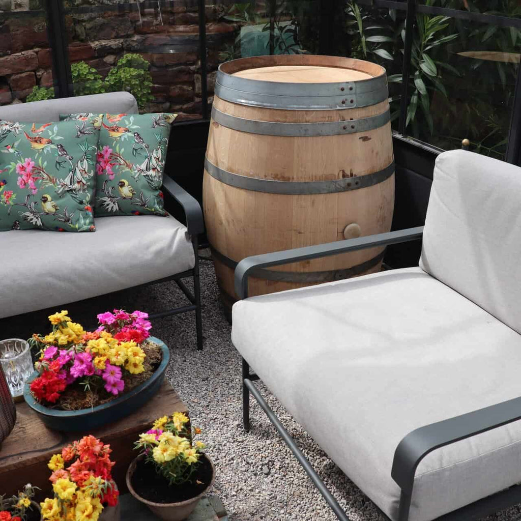 Eine gemütliche Sitzecke auf der Terrasse mit zwei gepolsterten Stühlen, bunten Blumenkissen, einem Holztisch, bunten Blumen und einem Original Weinfass aus Eiche, Barriquefass 225 Liter im Hintergrund, alles eingebettet in üppiges Grün.