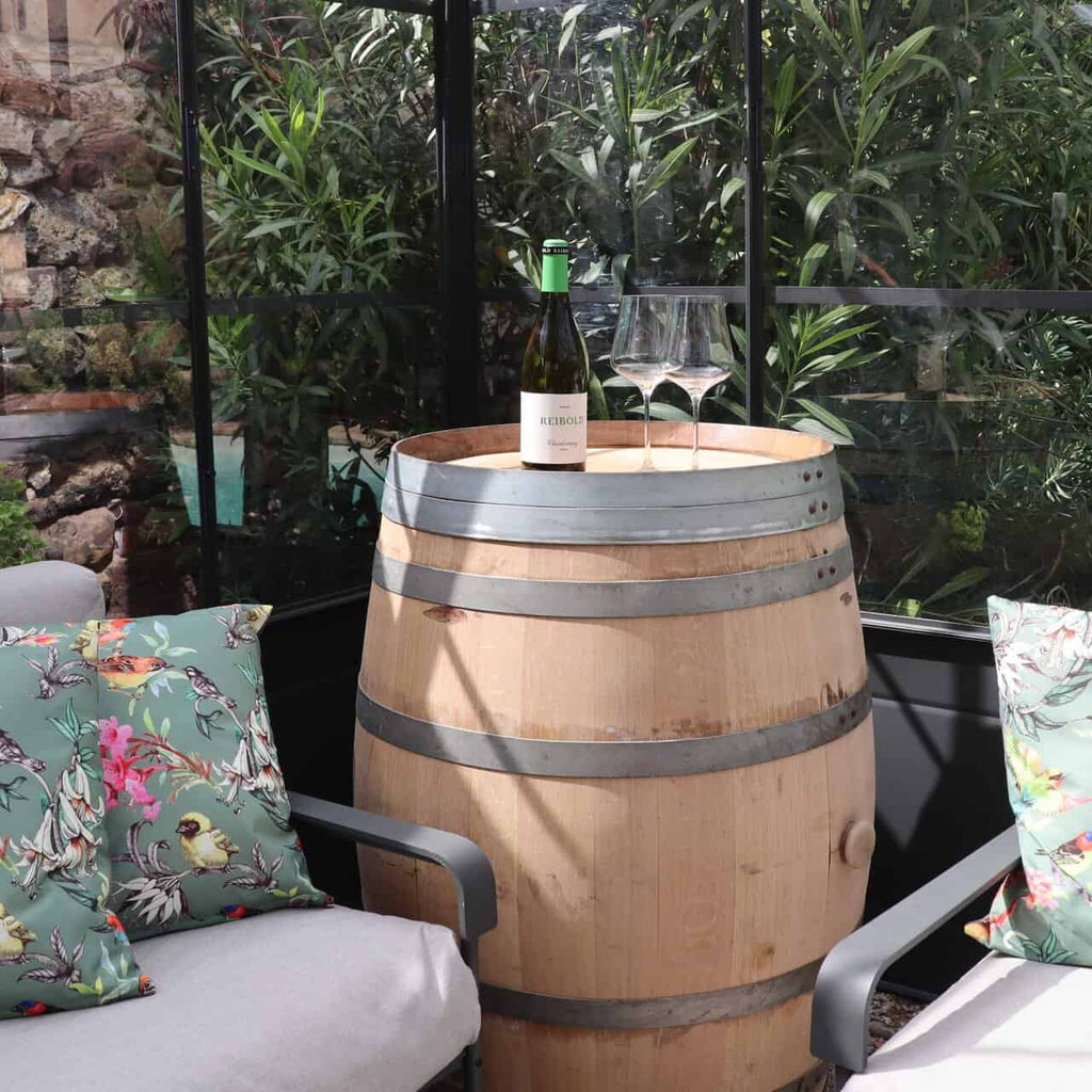 Ein Original Weinfass aus Eiche, Barriquefass 225 Liter, hält eine Flasche Weißwein und zwei leere Gläser, umringt von gepolsterten Terrassenstühlen mit Blumenkissen in einem sonnenbeschienenen Raum mit Blattpflanzen, die durch das Fenster zu sehen sind.