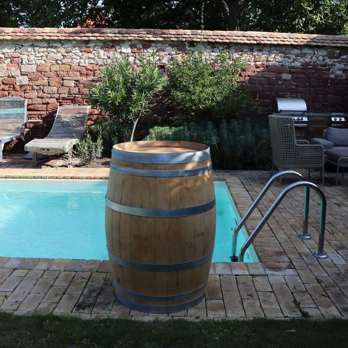 Ein Original Weinfass aus Eiche, Barriquefass 225 Liter, steht neben einem strahlend blauen Pool, umgeben von einer Backsteinmauer. Liegestühle, ein Grill und Sitzgelegenheiten auf der Terrasse mit Steinfliesen runden das Bild ab.