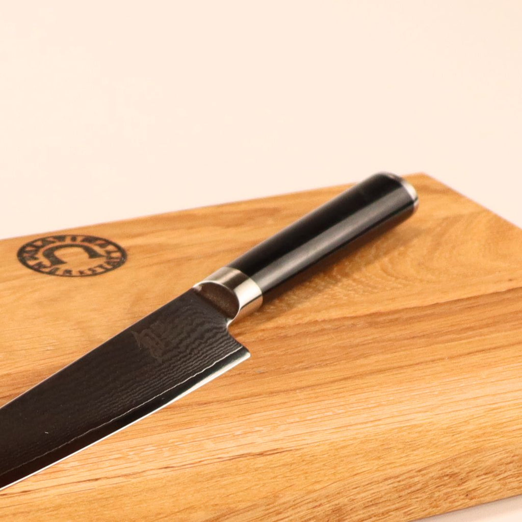 Ein Kai Shun DM-0701 Allzweckmesser mit 15 cm Klinge und schwarzem Griff liegt auf einem 30x20 cm Schneidebrett aus Eiche mit rundem Logo in der Ecke, vor hellem Hintergrund.