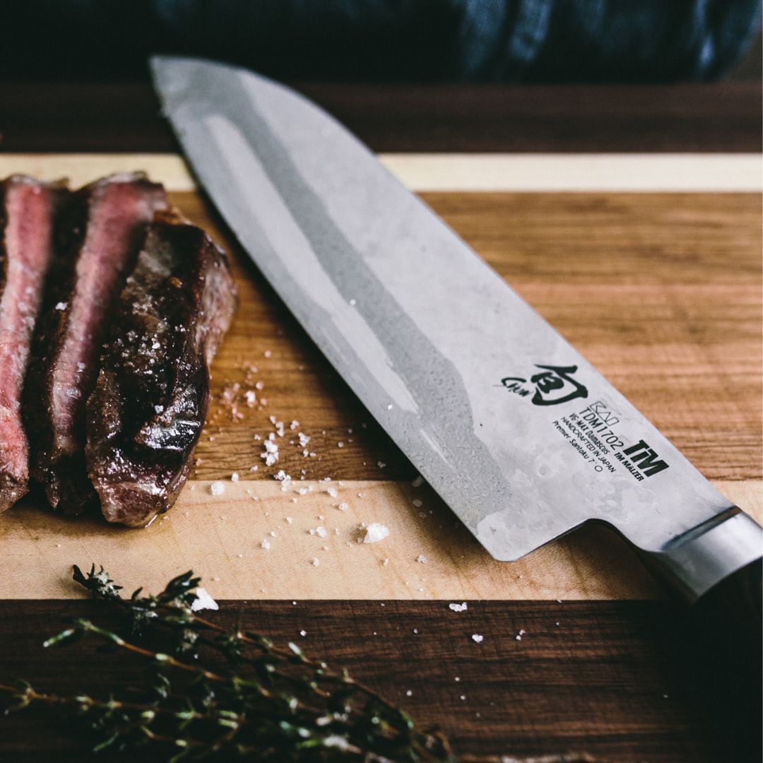 Ein Kai Shun Tim Mälzer Santoku TDM-1702 mit einer 18 cm langen Klinge aus "THE MAGIC KAI WISHLIST" liegt auf einem Holzbrett neben drei medium-rare Steakscheiben, Salz und frischen Kräutern.