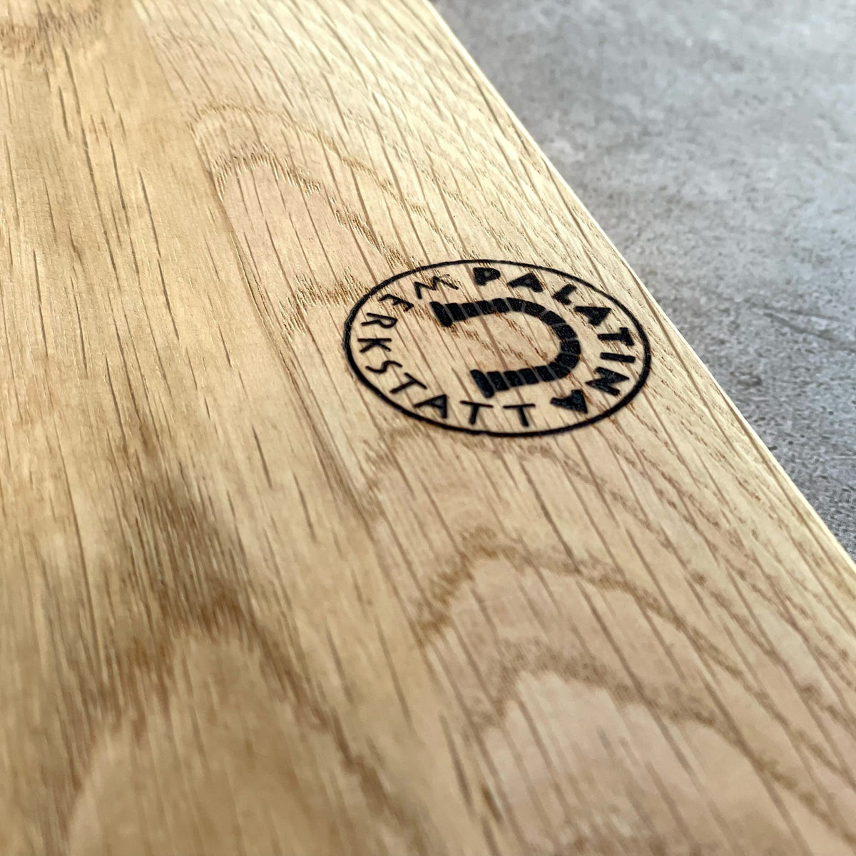 Nahaufnahme des Schneidebretts aus Eichenholz (30x20 cm) mit kreisförmigem "PALATIN WERKSTATT"-Logo und Hufeisenmotiv, das im Set KAI Shun Nagare NDC-0702 Santoku-Messer 18cm enthalten ist. Die elegante Holzmaserung ist deutlich sichtbar.