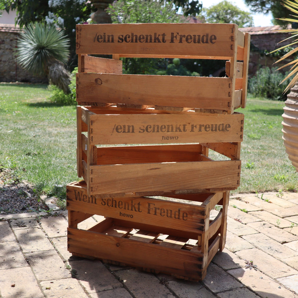 Drei „Alte Weinkisten Holz Vintage aus der Pfalz mit Aufschrift“ sind in einem Garten mit Ziegelweg und grünem Rasen gestapelt. Sonnenlicht wirft Schatten und hebt die rustikale Textur dieser charmanten Kisten und der umgebenden Pflanzen hervor.