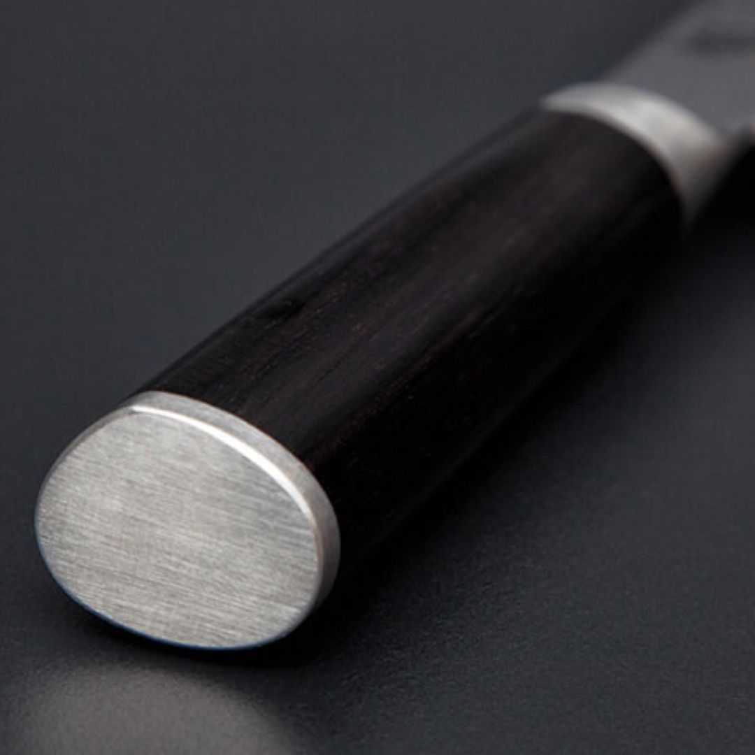 Nahaufnahme des abgerundeten Metallendes eines dunklen Holzmessergriffs aus dem KAI Shun Classic Set DM-W25, Allzweckmesser+Santoku, "THE MAGIC KAI WISHLIST", der auf einer glatten, dunklen Oberfläche ruht.