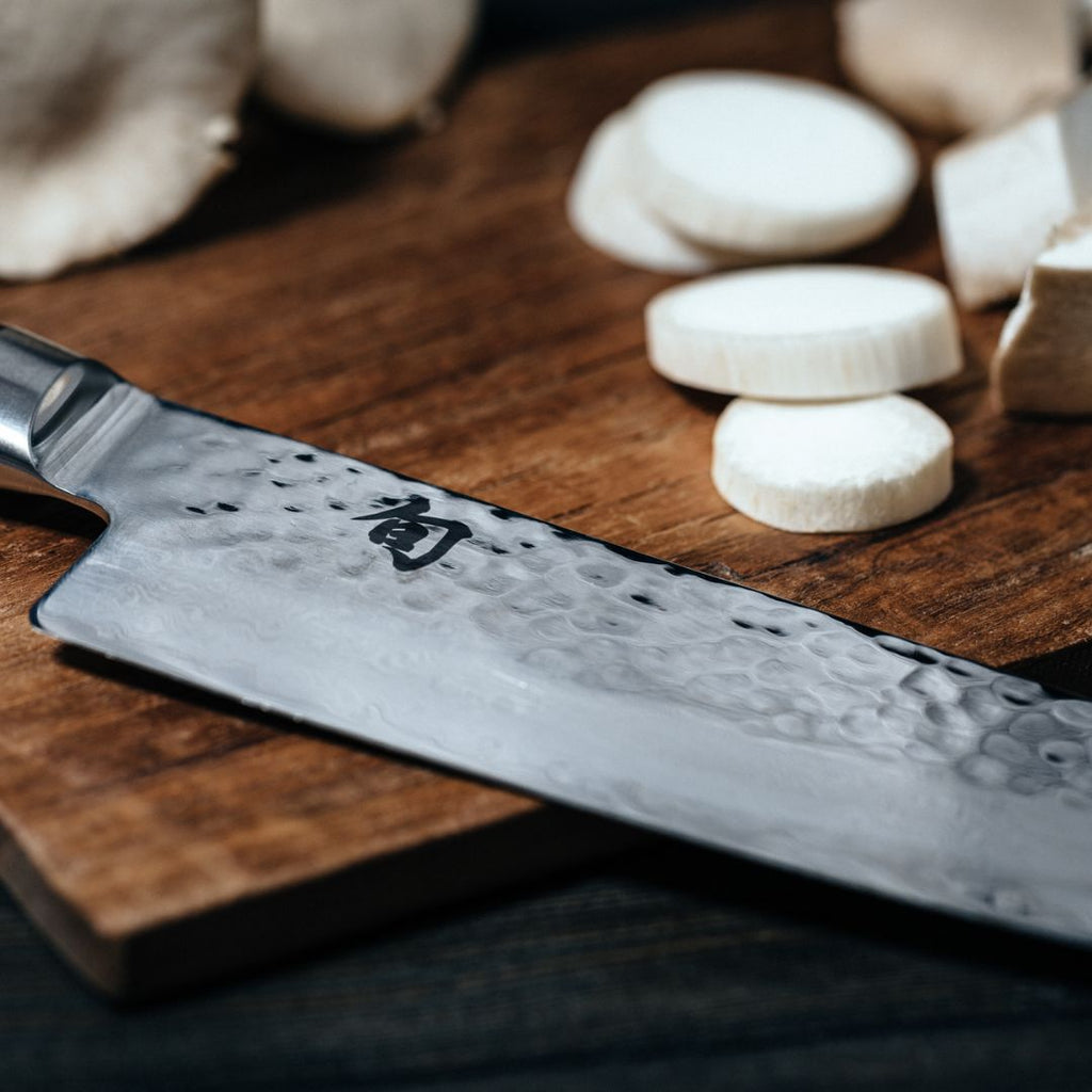 Eine Nahaufnahme des Kai Shun Tim Mälzer Santoku TDM-1702 (18 cm) "THE MAGIC KAI WISHLIST", dessen gehämmerte Klinge mit japanischen Kanji auf einem hölzernen Schneidebrett neben geschnittenem weißen Gemüse liegt.