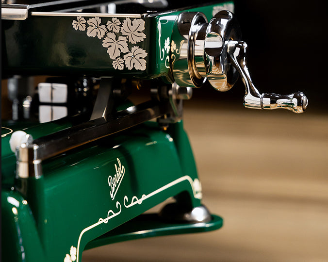 Nahaufnahme einer grünen Berkel Volano B114 Sondermodell Schwungradmaschine incl. Standfuß + Fassholzbrett, mit silbernen Blumendetails und verchromter Handkurbel, vor einem hellbraunen Hintergrund. Das "Berkel"-Logo ist auf dem Sockel sichtbar.