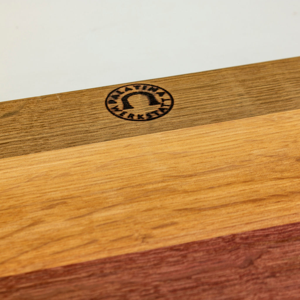 Nahaufnahme eines handgefertigten Holzschneidebretts mit den Maßen 40 x 30 cm, das vier verschiedene Holzmaserungen und Farben aufweist. In der oberen linken Ecke befindet sich eine kreisförmige Gravur mit dem Text „Klabauter Werkstatt“ und einem Bogensymbol innerhalb des Kreises. Das elegante Kai Shun Classic White | DM-0705W Brotmesser mit einer 23 cm langen Damastklinge steht anmutig vor dem cremefarbenen Oberflächenhintergrund.