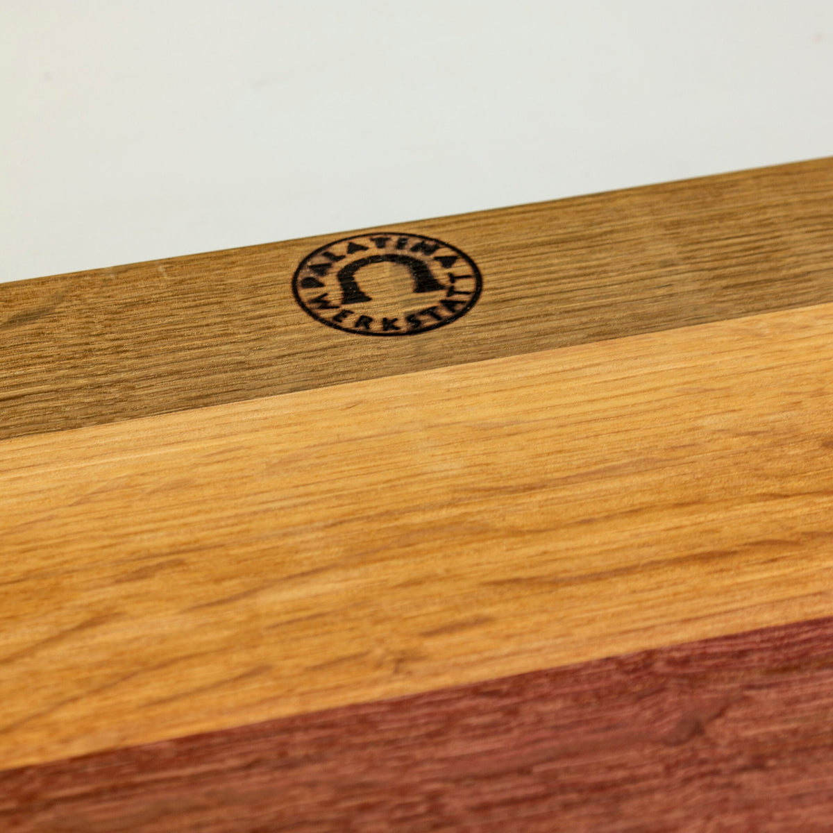 Nahaufnahme eines handgefertigten Holzschneidebretts mit den Maßen 40 x 30 cm, das vier verschiedene Holzmaserungen und Farben aufweist. In der oberen linken Ecke befindet sich eine kreisförmige Gravur mit dem Text „Klabauter Werkstatt“ und einem Bogensymbol innerhalb des Kreises. Das elegante Kai Shun Classic White | DM-0705W Brotmesser mit einer 23 cm langen Damastklinge steht anmutig vor dem cremefarbenen Oberflächenhintergrund.