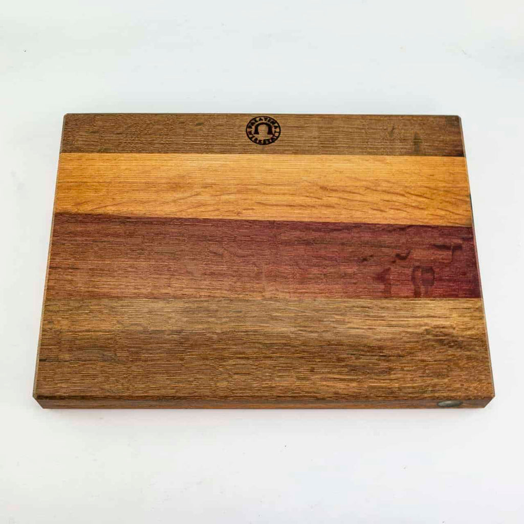 Ein rechteckiges Schneidebrett aus Holz mit verschiedenen Holzmaserungen, mit hellen, mittleren und dunklen Holzstreifen für einen mehrfarbigen Effekt. In die obere Mittelkante des Schneidebretts ist ein kleines, rundes Logo eingraviert. Das Brett liegt auf einem schlichten weißen Hintergrund.