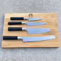 Das Kai Wasabi Black Kochmesser-Set, bestehend aus vier ultra-scharfen Messern mit schwarzen Griffen – einem Schälmesser, einem Universalmesser, einem Santokumesser und einem Brotmesser mit Wellenschliff – wird auf einem großen 40x25 cm großen Küchenbrett aus Eiche präsentiert, das oben in der Mitte mit einem Logo versehen ist.