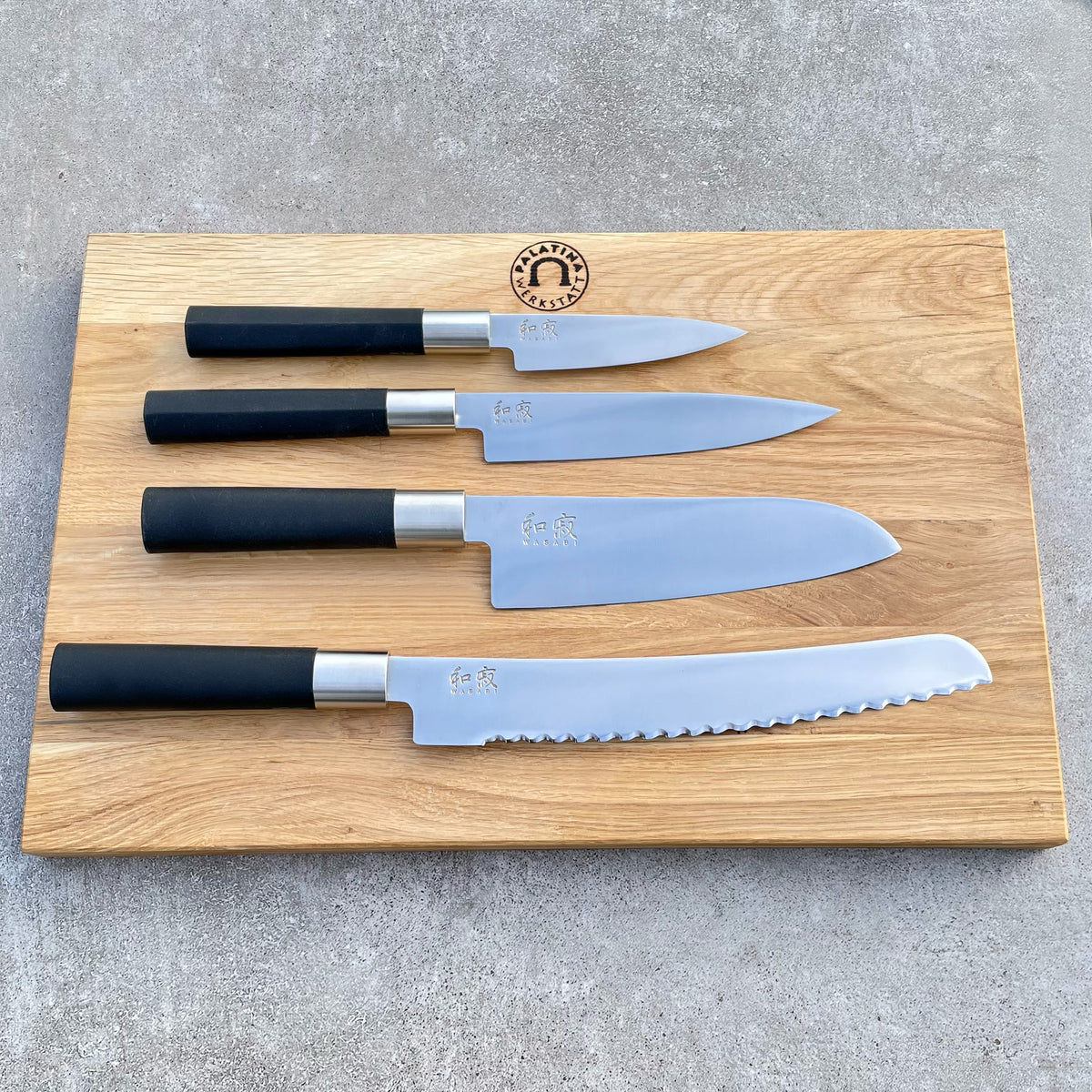 Das Kai Wasabi Black Kochmesser-Set, bestehend aus vier ultra-scharfen Messern mit schwarzen Griffen – einem Schälmesser, einem Universalmesser, einem Santokumesser und einem Brotmesser mit Wellenschliff – wird auf einem großen 40x25 cm großen Küchenbrett aus Eiche präsentiert, das oben in der Mitte mit einem Logo versehen ist.