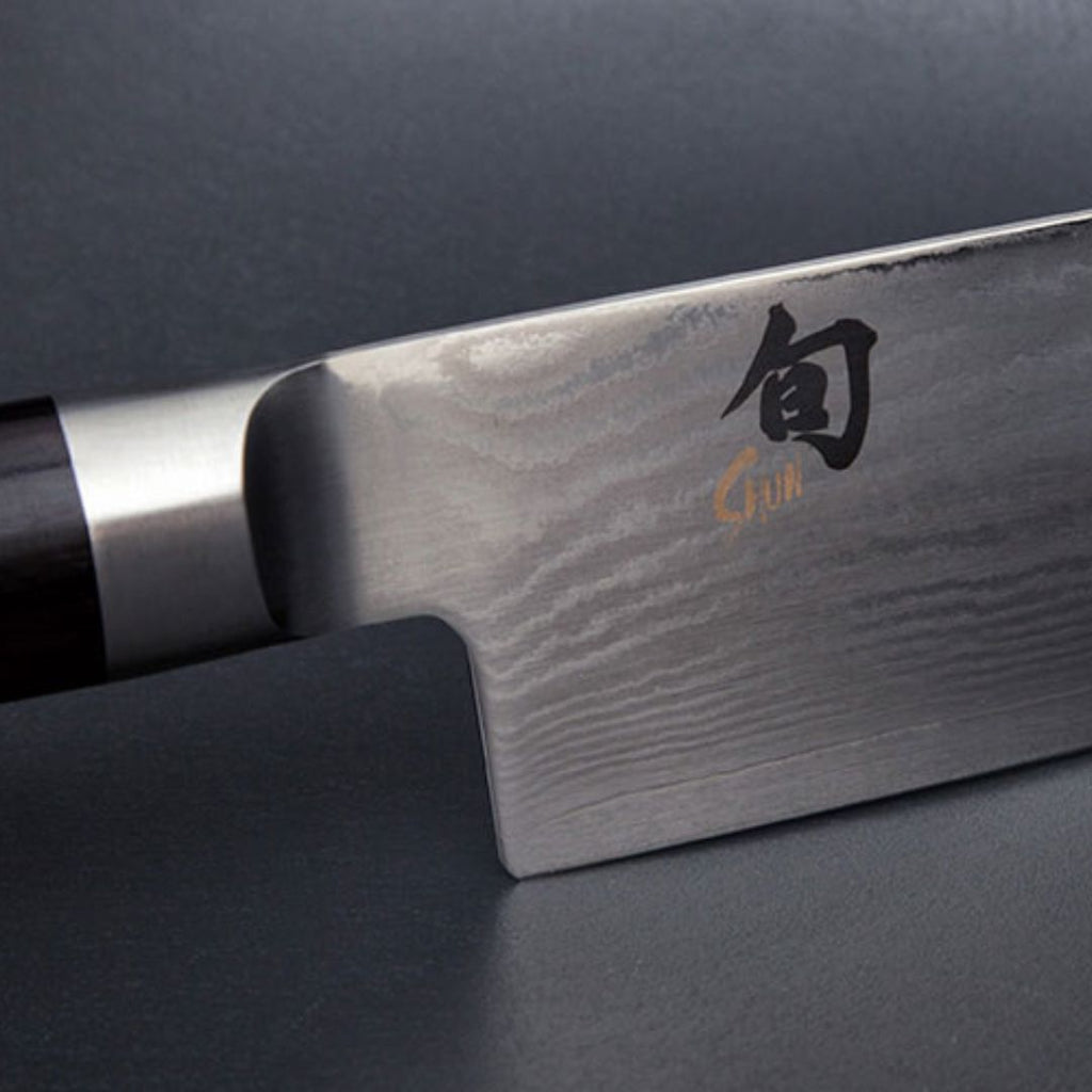 Nahaufnahme eines KAI Shun Classic Set DM-W25 Allzweckmesser+Santoku aus "THE MAGIC KAI WISHLIST", mit dunklem Griff und Klinge aus Damaszenerstahl mit Wellenmustern und japanischen Schriftzeichen vor einem glatten, dunklen Hintergrund.