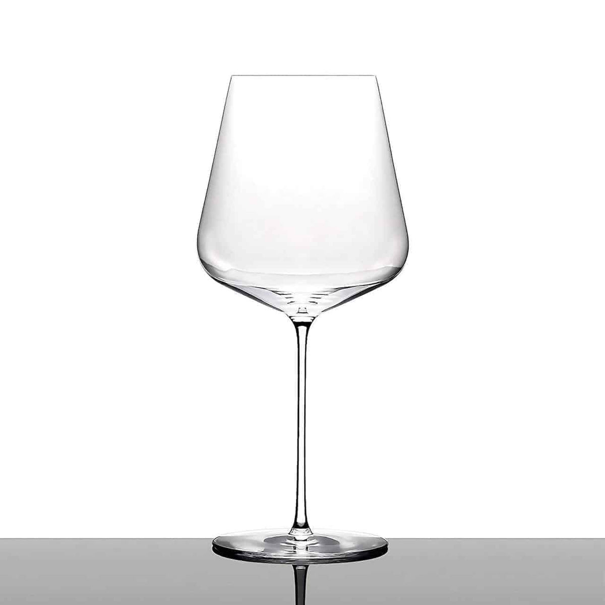 Ein klares, leeres mundgeblasenes Bordeaux-Weinglas von Zaltoglas Glasmanufaktur Denk´Art mit dünnem Stiel und breiter Schale steht auf einer reflektierenden Oberfläche vor einem weißen Hintergrund. Das elegante Design zeichnet sich durch klare Linien und eine moderne Silhouette aus und unterstreicht die Schönheit dieser Kreation von Zaltoglas.